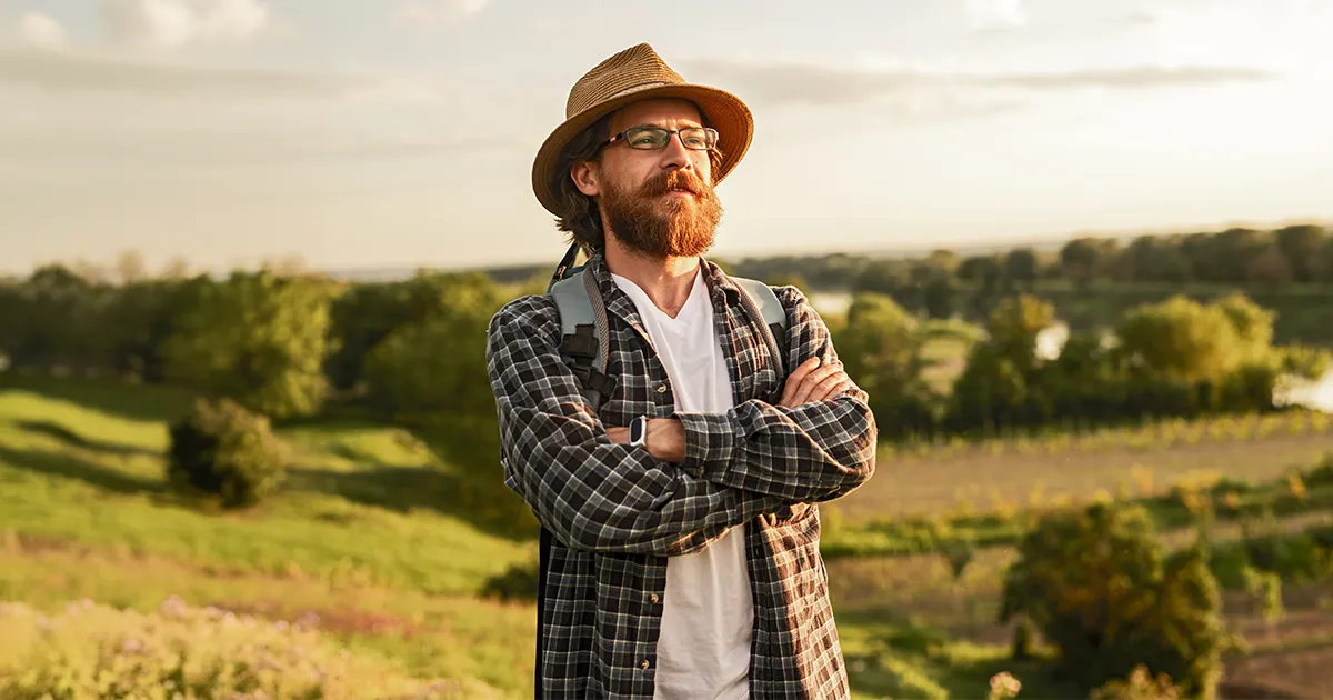 Homme avec un chapeau de paille, une barbe rousse portant un sac à dos dans une prairie.
