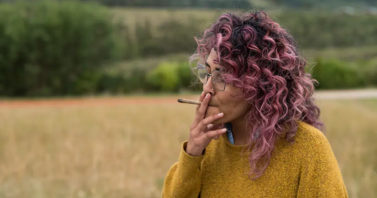 Femme au cheveux rose dans un champ entrain de fumer un joint de cannabis.